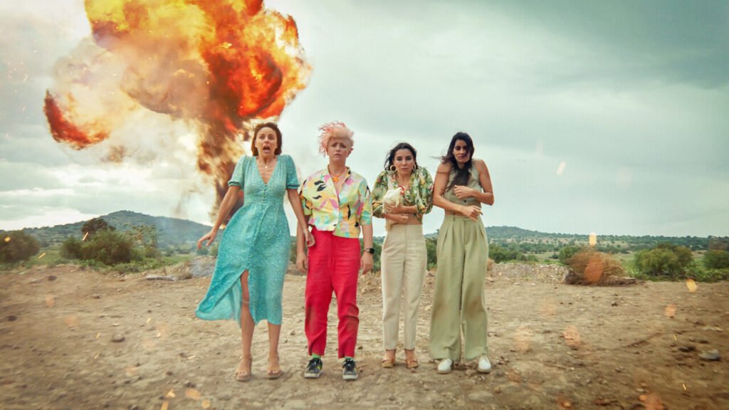 Queens on the run review - A still from 'Queens on the Run'. The cast is standing on a cliff and there is an explosion behind them.